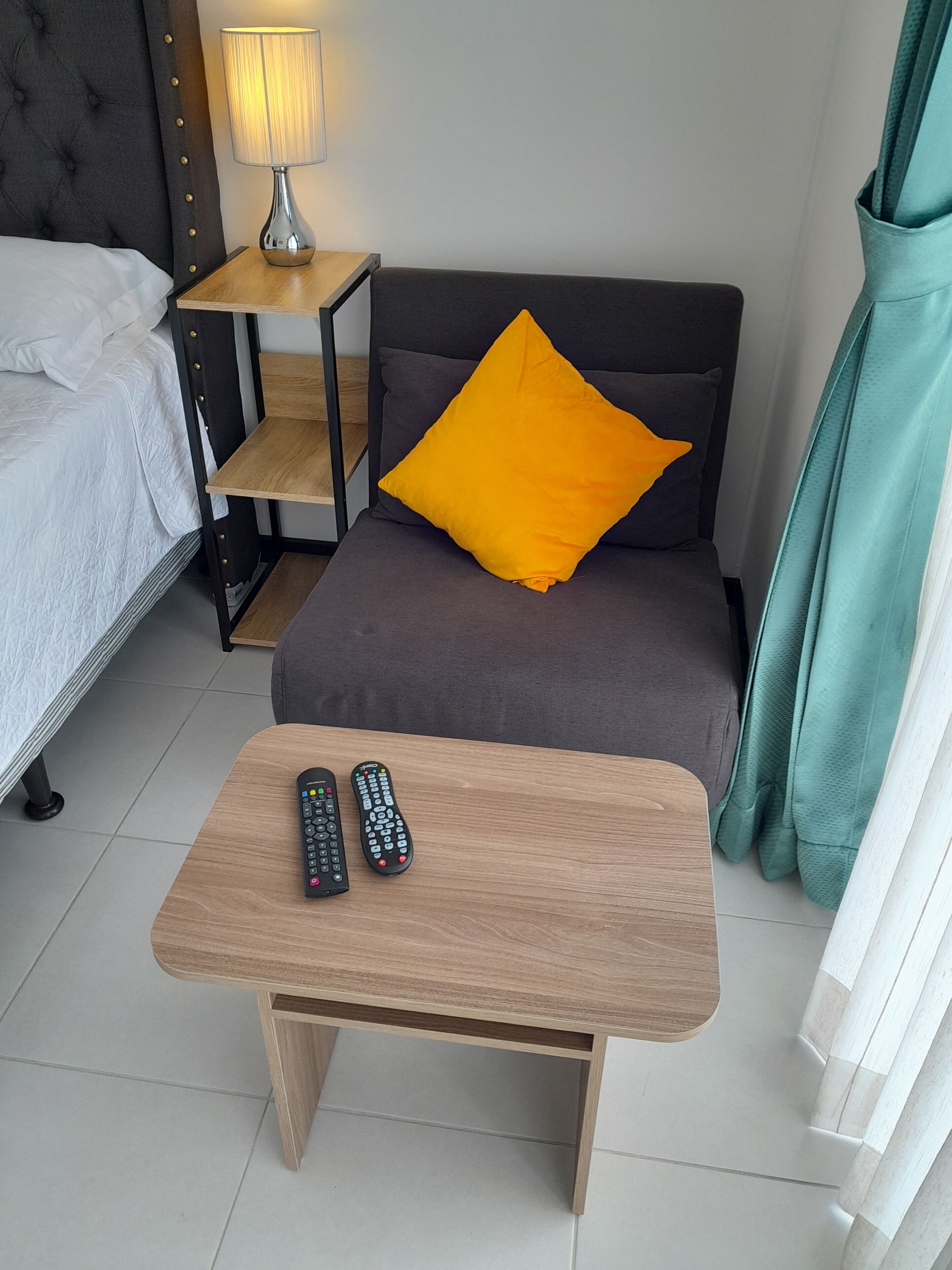 Une chambre d’hôtel avec un lit, un canapé agrémenté d’un coussin jaune, une petite table avec deux télécommandes, une lampe et un rideau.