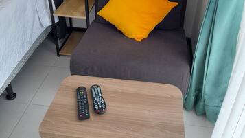 Une chambre d’hôtel avec un lit, un canapé agrémenté d’un coussin jaune, une petite table avec deux télécommandes, une lampe et un rideau.