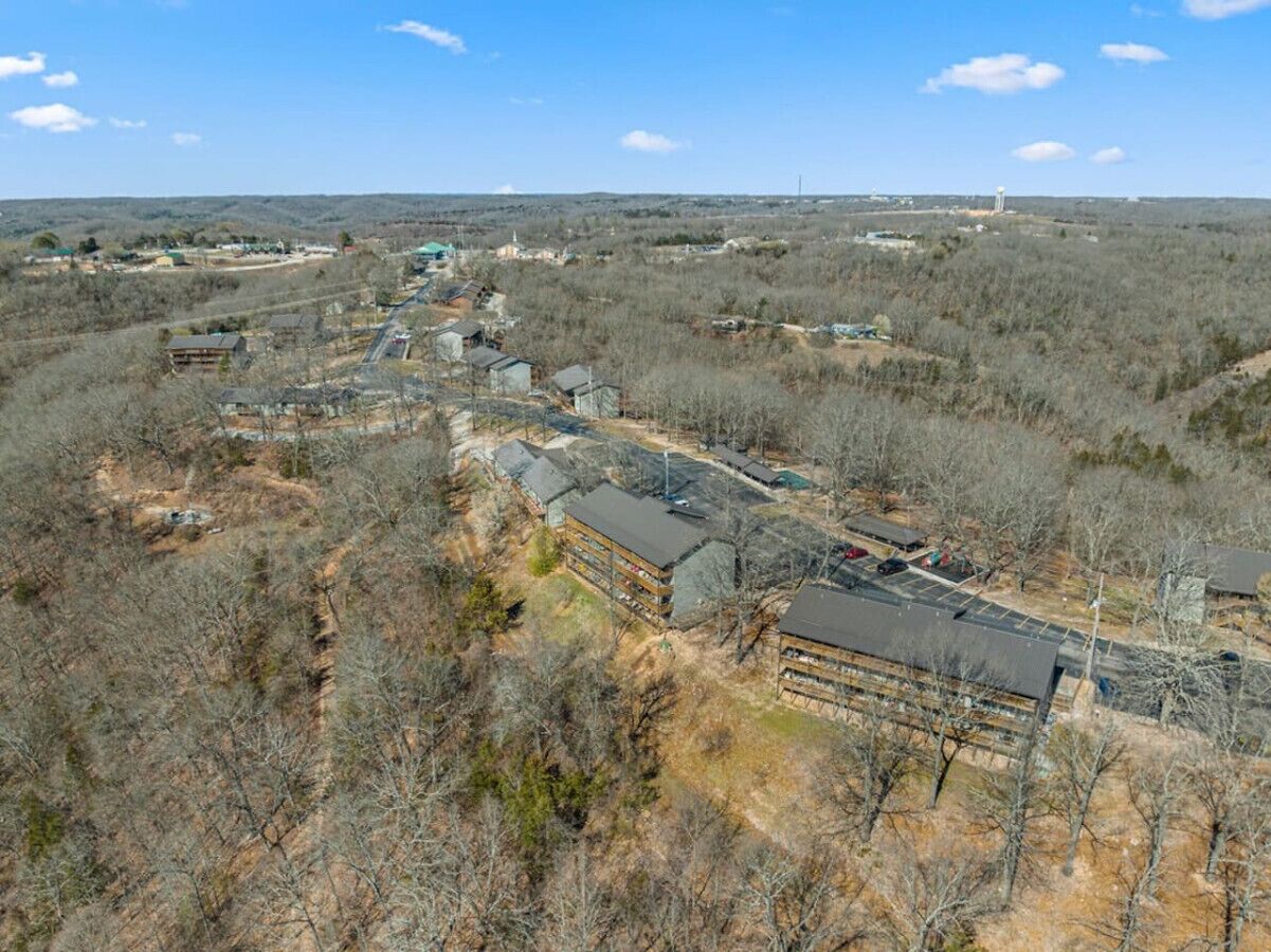 An aerial view of a residential area with multiple buildings, including a large two-story house with a garage, surrounded by trees and open land.