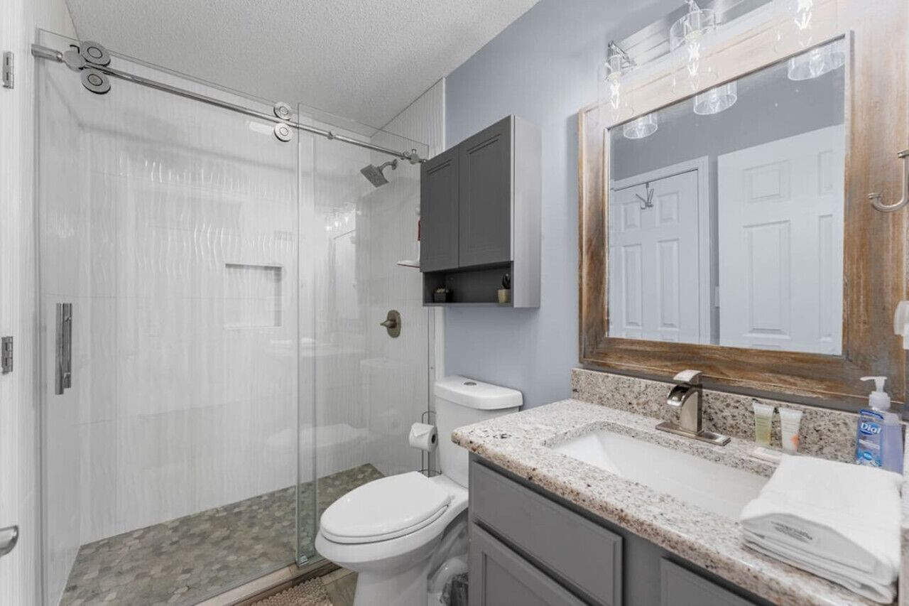 A modern bathroom with a glass shower enclosure, a white toilet, a granite countertop sink, and a large mirror with a wooden frame.