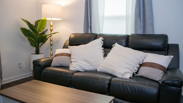 Een zwarte leren bank met witte en gestreepte kussens, een houten salontafel, een potplant en een vloerlamp in een moderne woonkamer met raam en gordijnen.