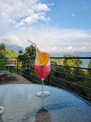 Un vaso de bebida rosa con una rodaja de naranja sobre una mesa, con vistas a montañas y vegetación.