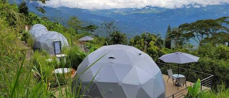 A geodesic dome structure surrounded by lush greenery, with a view of distant mountains and a cloudy sky.