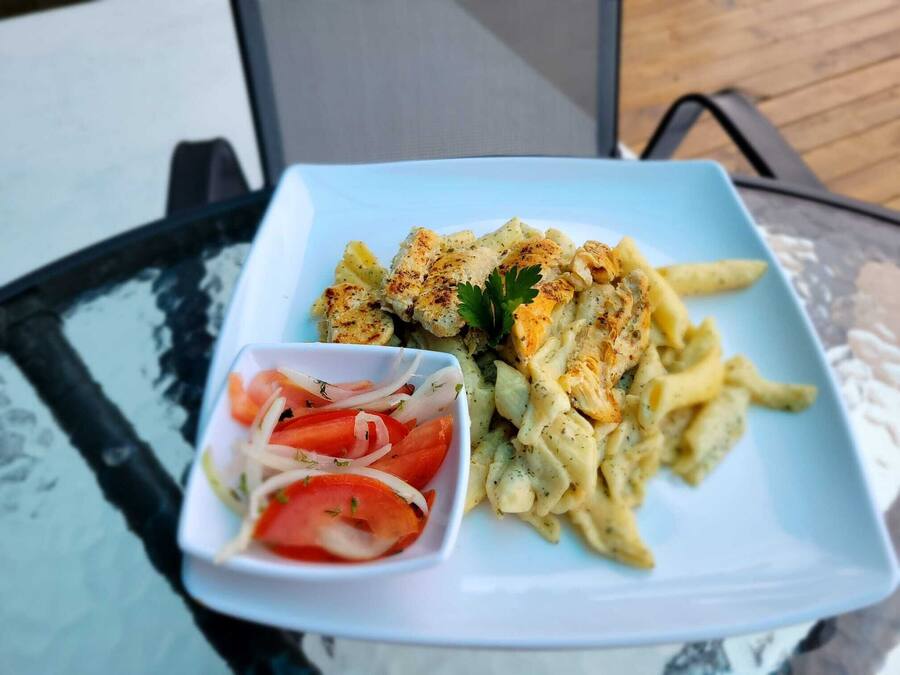 A plate of pasta with chicken and a small bowl of tomato salad on a glass table.