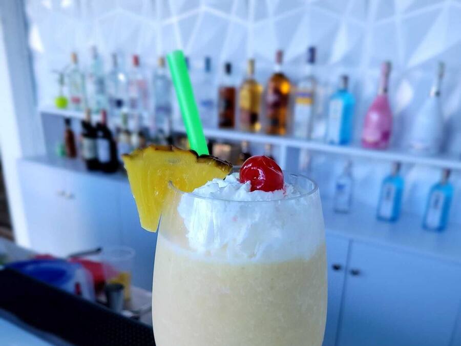 Cóctel con una rodaja de piña y una cereza en un vaso, frente a una barra con varias botellas al fondo.