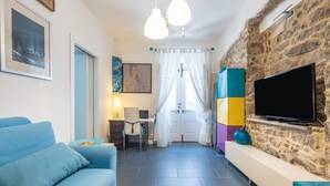 A living room with a blue sofa, a TV mounted on a stone wall, a desk with a chair, and a door with a curtain. - Apartment 'Casa Nora' with Private Terrace, Wi-Fi and Air Conditioning (Calasetta)