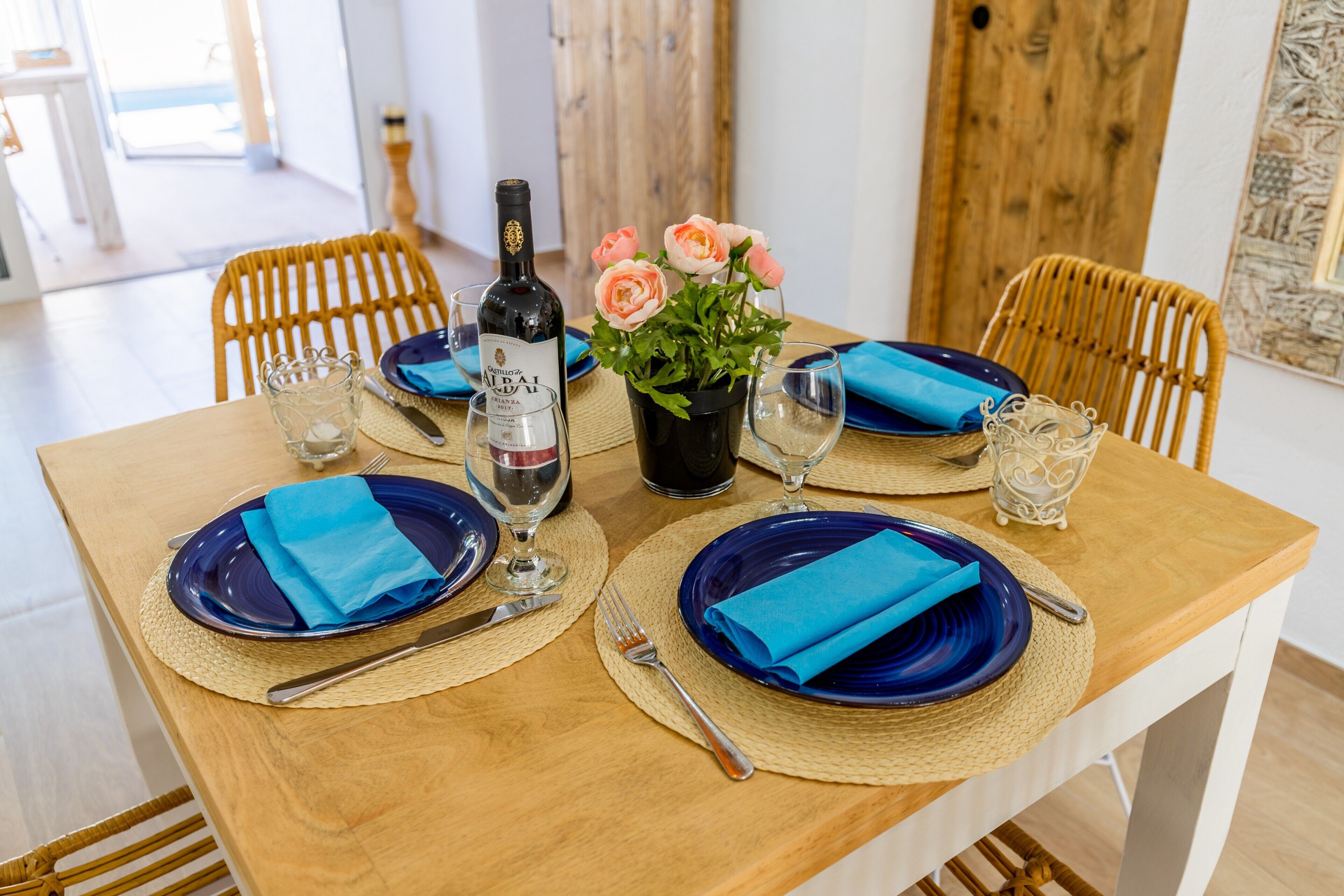 Una mesa de comedor dispuesta con platos azules, servilletas y una botella de vino, rodeada de sillas de mimbre.