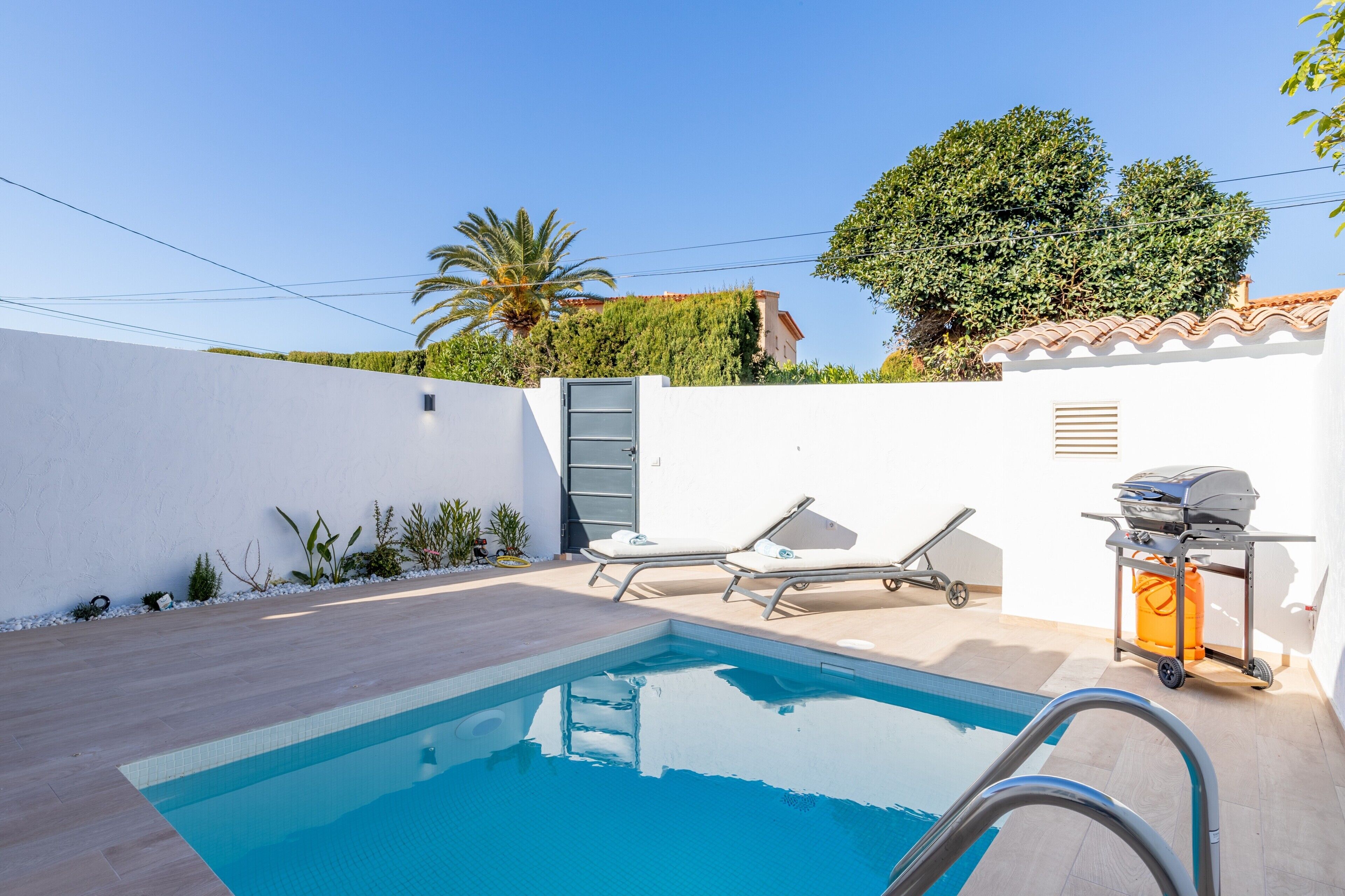 A swimming pool with lounge chairs, a grill cart, and a white wall with a door.