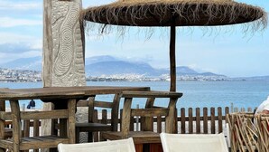 Un espace extérieur aménagé avec des sièges en bois et un parasol en chaume, offrant une vue sur une étendue d’eau et sur le paysage urbain au loin.