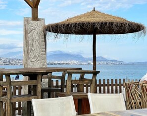 Un espace extérieur aménagé avec des sièges en bois et un parasol en chaume, offrant une vue sur une étendue d’eau et sur le paysage urbain au loin.