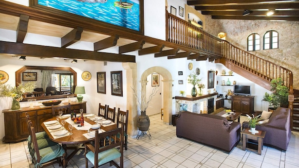 A spacious living area with a high ceiling, a dining table set for four, a staircase with wooden railings, and a stone wall.