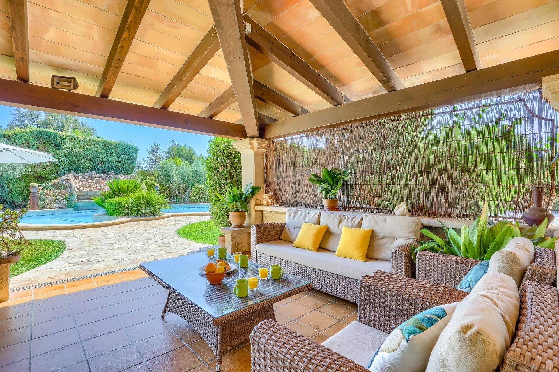 Un'area salotto esterna riparata con mobili in vimini, un tavolo in vetro e vista su un giardino curato e una piscina.