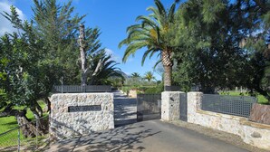 A gated entrance with stone walls and metal grilles, leading into a landscaped area with trees and a clear sky.