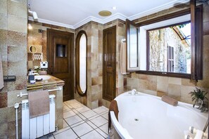 Une salle de bain avec une grande baignoire, un miroir, un lavabo avec des articles de toilette et une fenêtre donnant sur un mur en pierre.