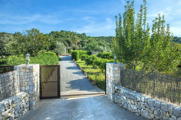Un chemin de pierres mène à un portail flanqué de chaque côté d’une clôture en métal, le tout étant entouré d’une végétation luxuriante et d’arbres.
