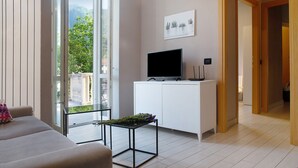 A modern living room with a sofa, a TV on a white cabinet, a glass table with flowers, and a view of greenery outside. - Apartment "Il Sole Di Calde Lago Maggiore 504" with Lake View, Balcony & Wi-Fi (castelveccana)