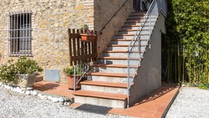 Edifici de pedra amb una escala que mena a una porta de fusta, envoltat de grava i plantes en testos.