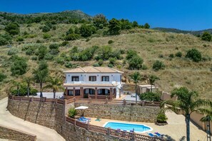 Une maison à deux étages avec une piscine, entourée d’un mur de pierre et de palmiers.
