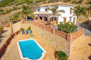 Une maison à deux étages avec une piscine, un patio doté de chaises longues et un mur en pierre.