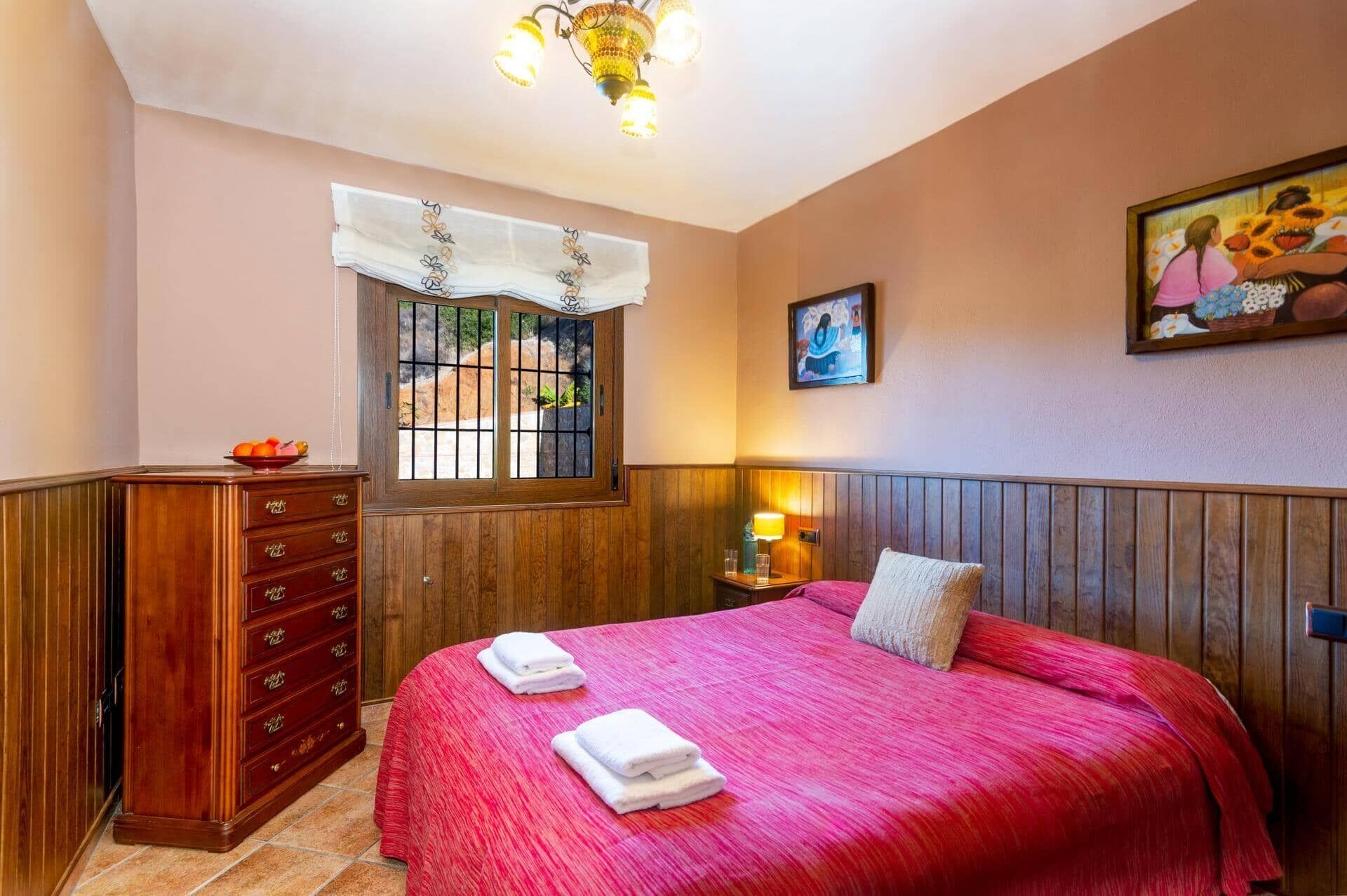 A hotel room with a bed, wooden dresser, window with bars, artwork on the wall, and a nightstand with a lamp.