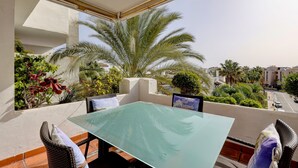 A balcony with a glass table, wicker chairs, and a view of palm trees and buildings. - Apartment Costalita with Sea View, Terrace, Shared Pool & Wi-Fi (Estepona)