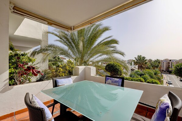 A balcony with a glass table, wicker chairs, and a view of palm trees and buildings.