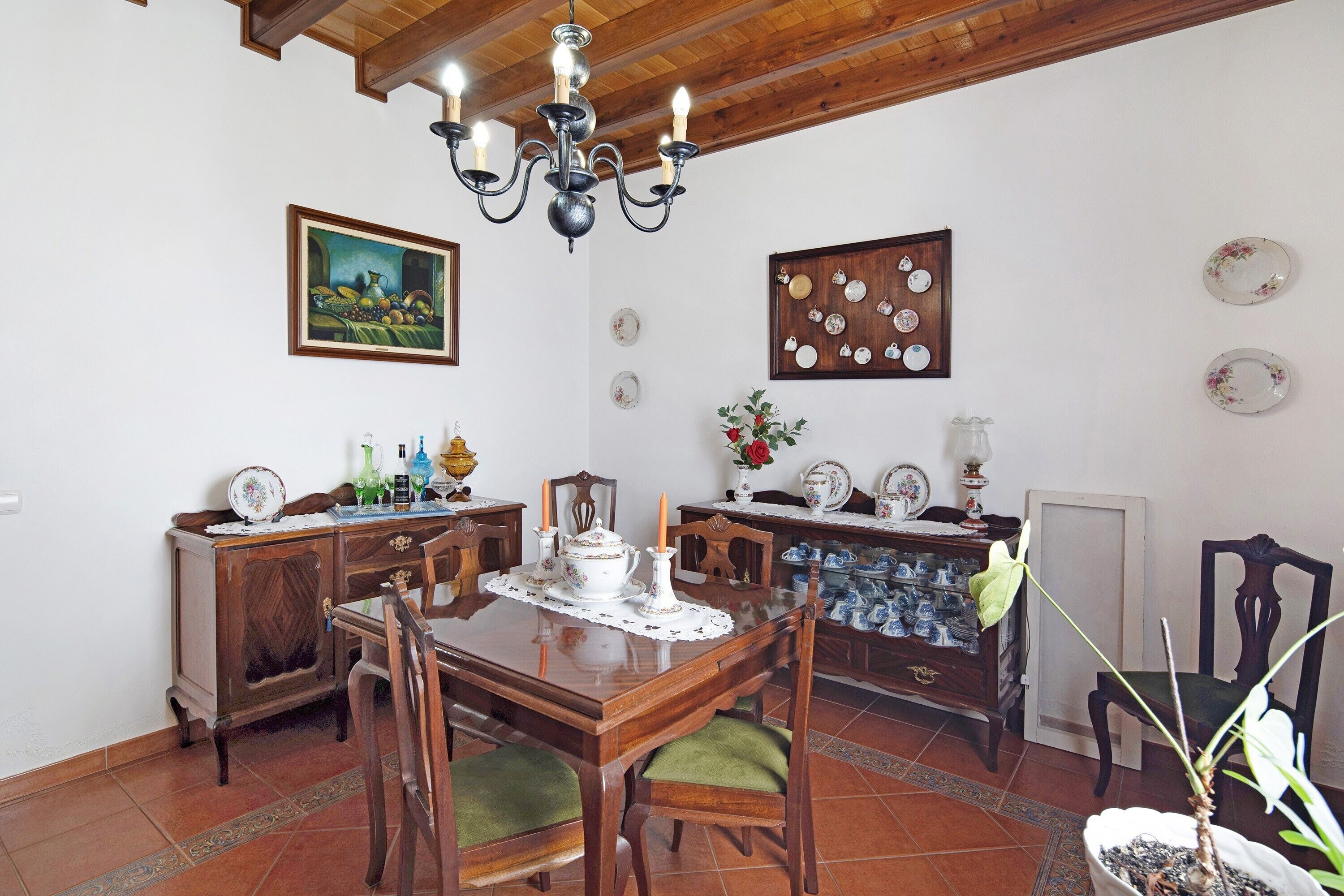 A dining area with a wooden table, chairs, a chandelier, a painting, and a wall-mounted plate display.