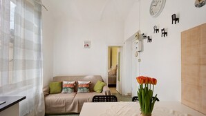 A room with a sofa, a table with a vase of flowers, a clock on the wall, and a tiled wall.
