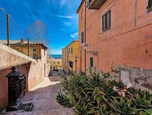 A narrow street with buildings on either side, a clear sky, and a view of the sea in the distance.