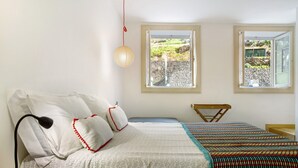 Ein Schlafzimmer mit Bett, Nachttisch, Lampe, zwei Fenstern und Blick auf einen Garten.