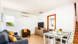 A living room with a sofa, a TV, a dining table with chairs, and a kitchen area with a cabinet and a window. - Villa Bini Mestral by Villa Plus (Binibeca)