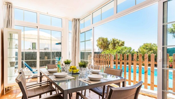 A dining area with a glass table and chairs, a view of a swimming pool, and a wooden fence. - Villa Bini Mestral by Villa Plus (Binibeca)