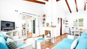 A spacious living area with a TV, wicker furniture, a fireplace, and a dining area with a view of the outdoors. - Villa Bini Anna by Villa Plus (Binibeca)