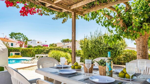 An outdoor dining area with a pergola, tables set for two, and a pool in the background. - Villa Bini Anna by Villa Plus (Binibeca)