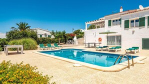 A pool area with lounge chairs and a table, surrounded by a white building with green shutters. - Villa Bini Anna by Villa Plus (Binibeca)