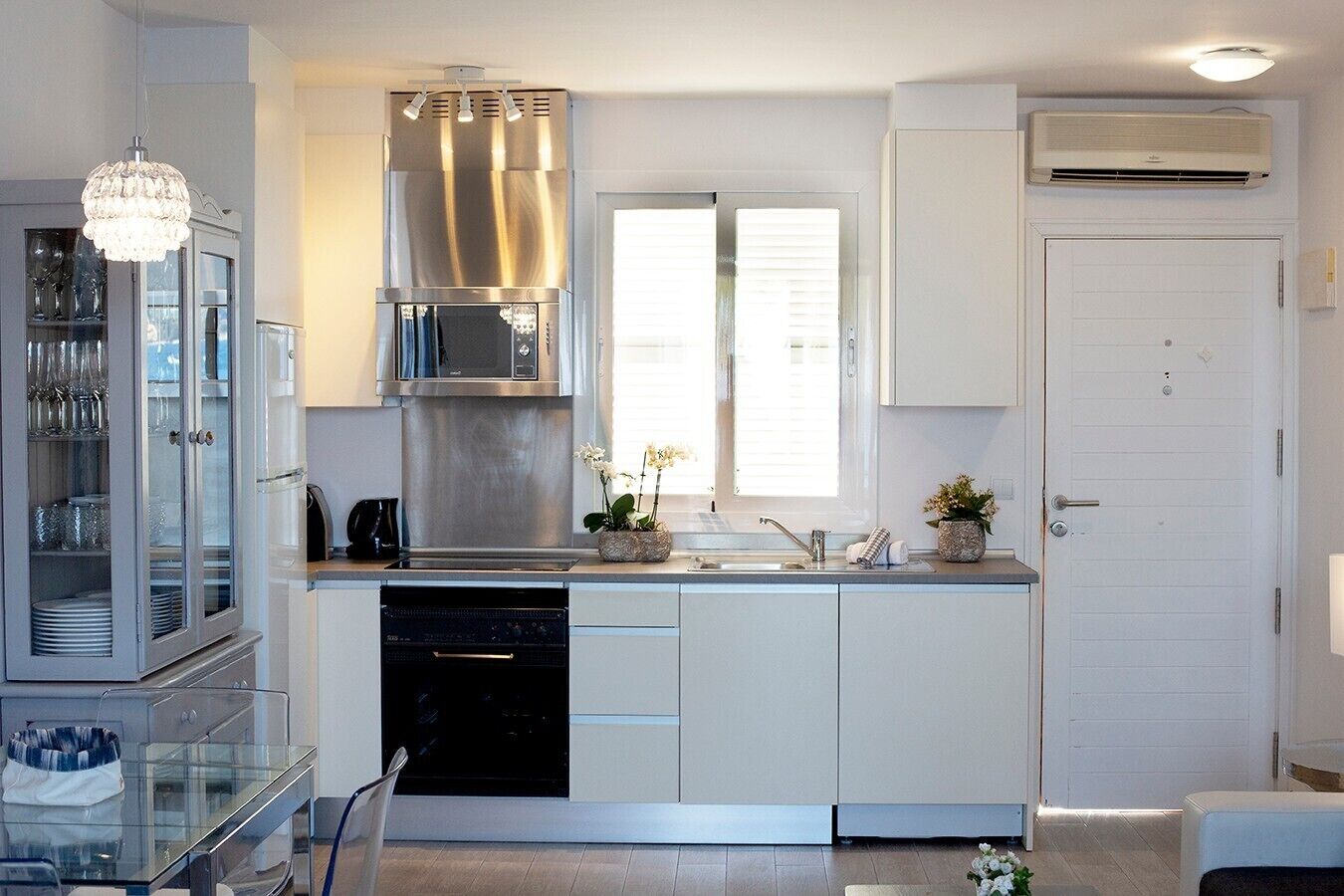 Ett modernt kök med en fläktkåpa i rostfritt stål, en diskmaskin och en inbyggd ugn. Det finns ett fönster med persienner, en krukväxt på bänkskivan och ett glasskåp med porslin.