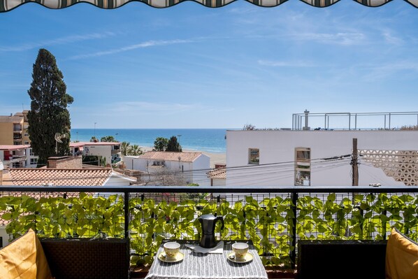 A balcony with a table set for two, a coffee pot, and a view of the sea and buildings.