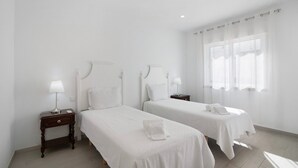 A hotel room with two beds, a nightstand with a lamp, and a window with sheer curtains.