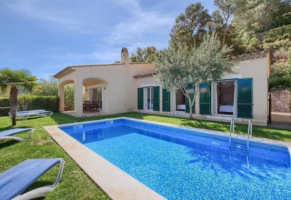 A swimming pool with lounge chairs, a covered patio, and a building with green shutters.