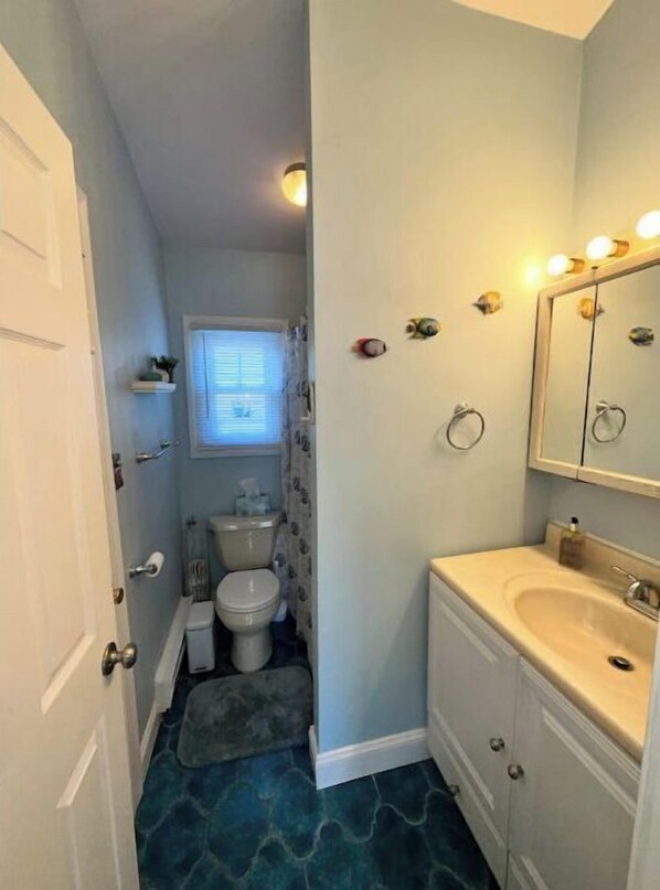 Baño con inodoro, lavabo y espejo. Luces decorativas en la pared y una ventana con persianas.