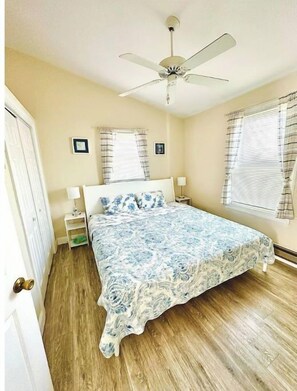 Une chambre à coucher comprenant un lit, un ventilateur de plafond, deux tables de chevet et deux fenêtres avec des rideaux.