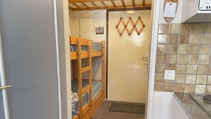 Een kleine keuken met een koelkast, een spoelbak en een klein aanrecht. In de aangrenzende kamer staat een houten stapelbed met een blauwe matras.