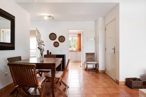 A dining area with a wooden table and chairs, a vase with dried flowers, and a kitchen in the background.