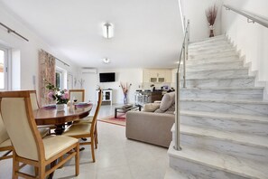 A modern living room with a dining area, a staircase, and a kitchen in the background.