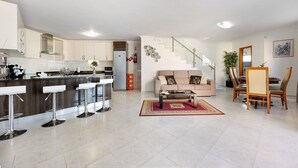 Une cuisine moderne avec un îlot central, un coin repas avec une table et des chaises en bois, un salon avec un canapé et une table basse, et un escalier menant à l’étage supérieur.