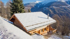 Um chalé de madeira com telhado coberto de neve, localizado numa encosta nevada com vista para as montanhas ao fundo.