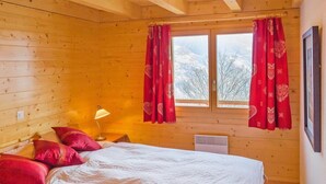 Quarto de cabana de madeira aconchegante, com cama, cortinas vermelhas e janela com vista para a neve.