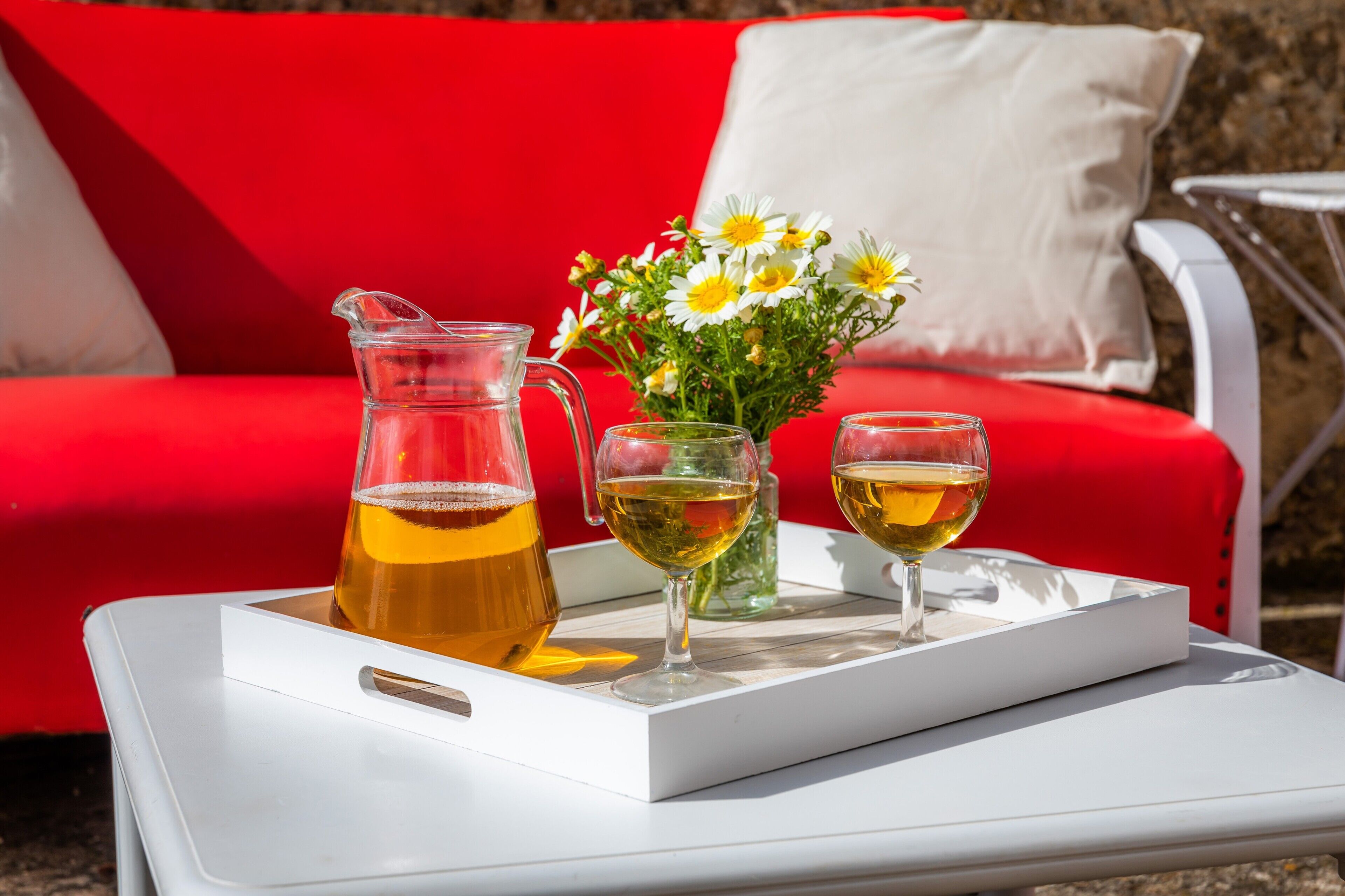 A tray with a pitcher and two glasses of a yellow beverage, a vase with white and yellow flowers, and a red couch in the background.