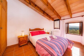 A bedroom with a wooden ceiling, a bed with a striped blanket, a nightstand with a lamp, and a window with a view.