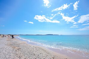 Ein Strand mit blauem Himmel, ruhigem Wasser, sandigem Ufer und sanften Hügeln in der Ferne.